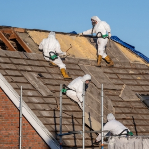 Sikringsarbejde ved nedtagning af asbesttag. Professionelle fagfolk i beskyttelsesudstyr fjerner gamle asbestholdige tagplader under sikre forhold.