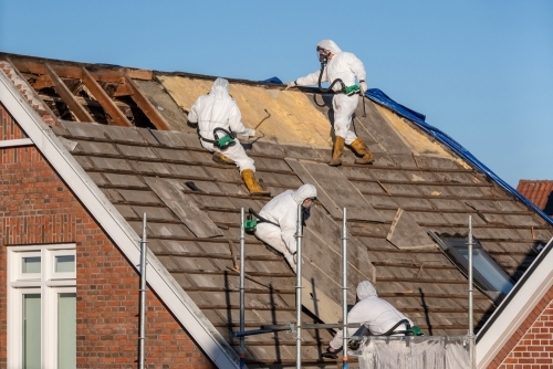 Sikringsarbejde ved nedtagning af asbesttag. Professionelle fagfolk i beskyttelsesudstyr fjerner gamle asbestholdige tagplader under sikre forhold.